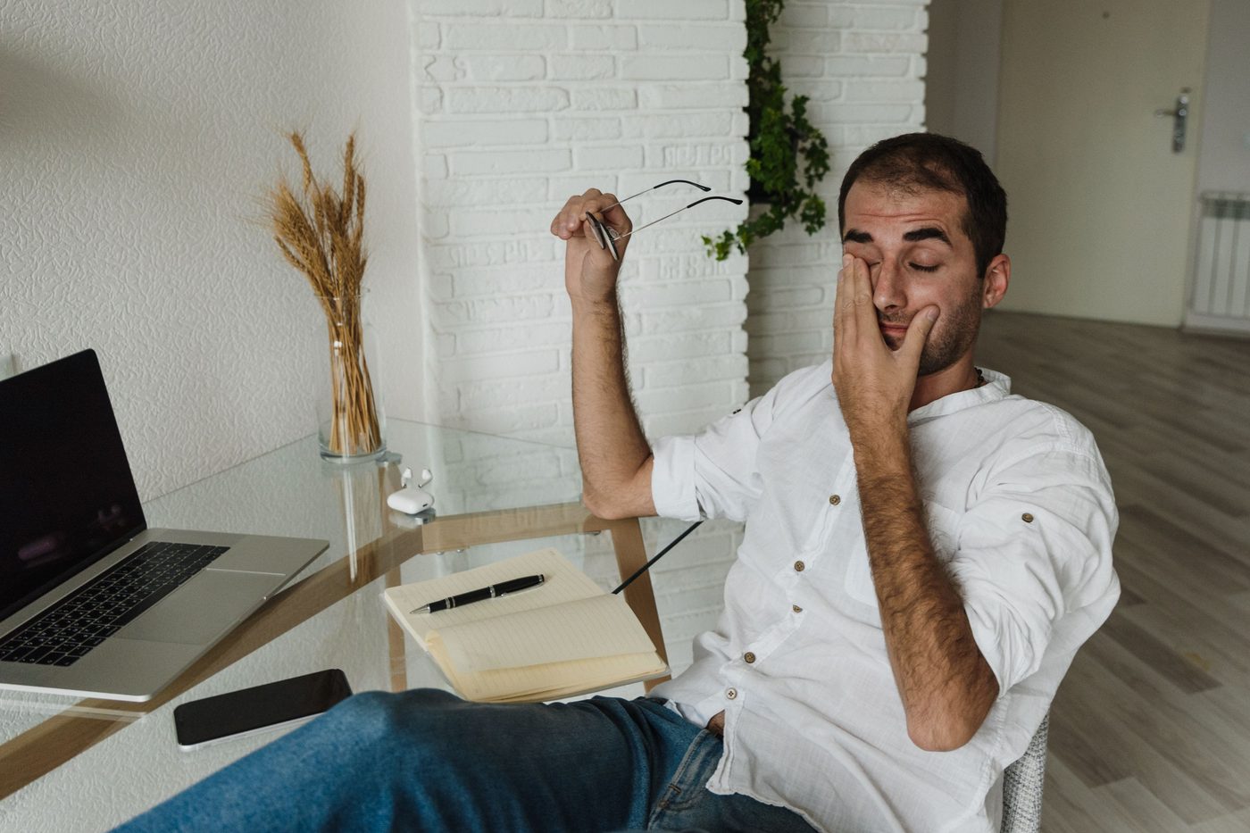 Man at a home office desk with a laptop, holding his glasses in one hand and rubbing his eyes with the other
