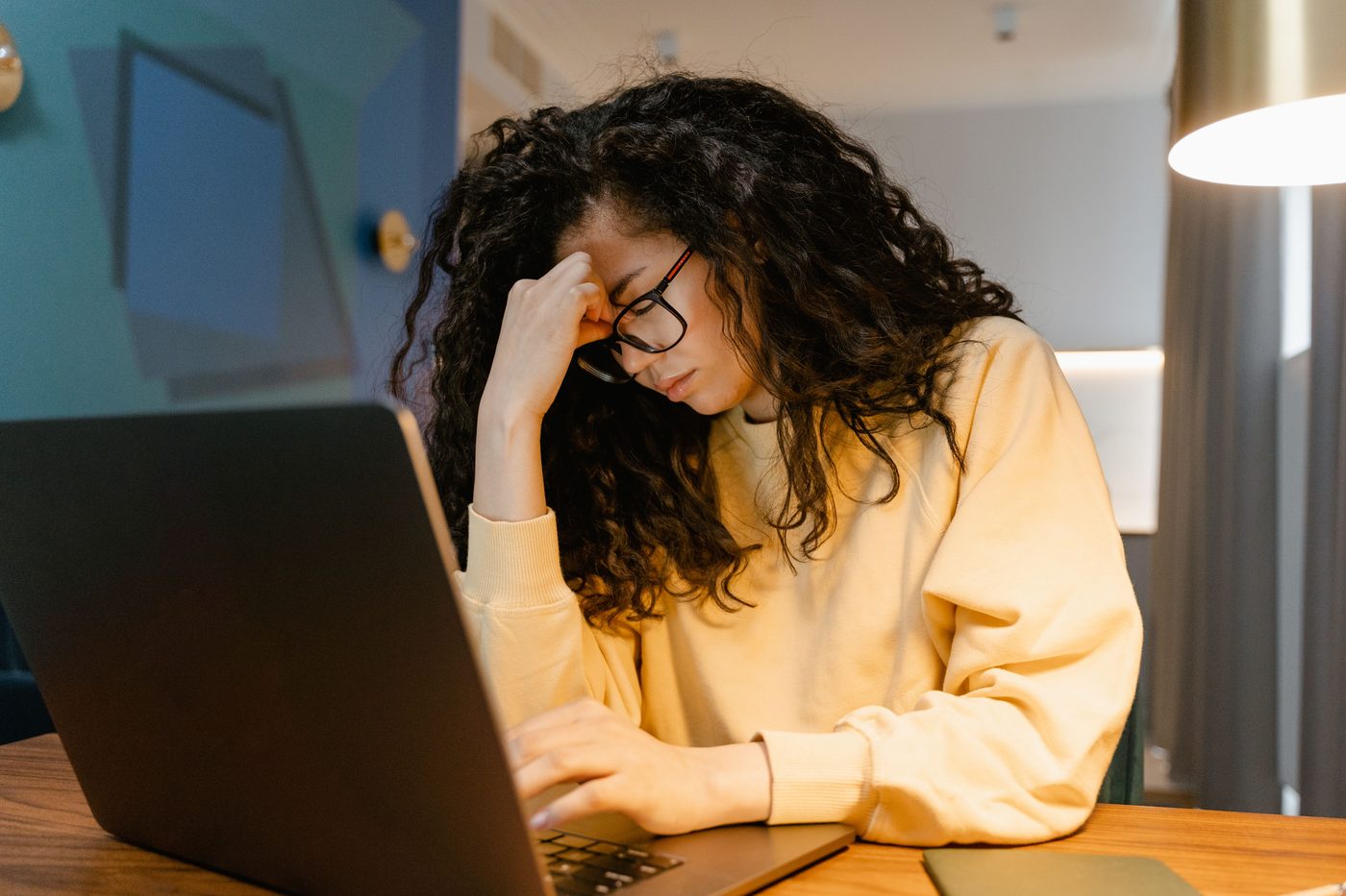 Woman with glasses at a laptop, eyes closed and hand pressed to her forehead in fatigue