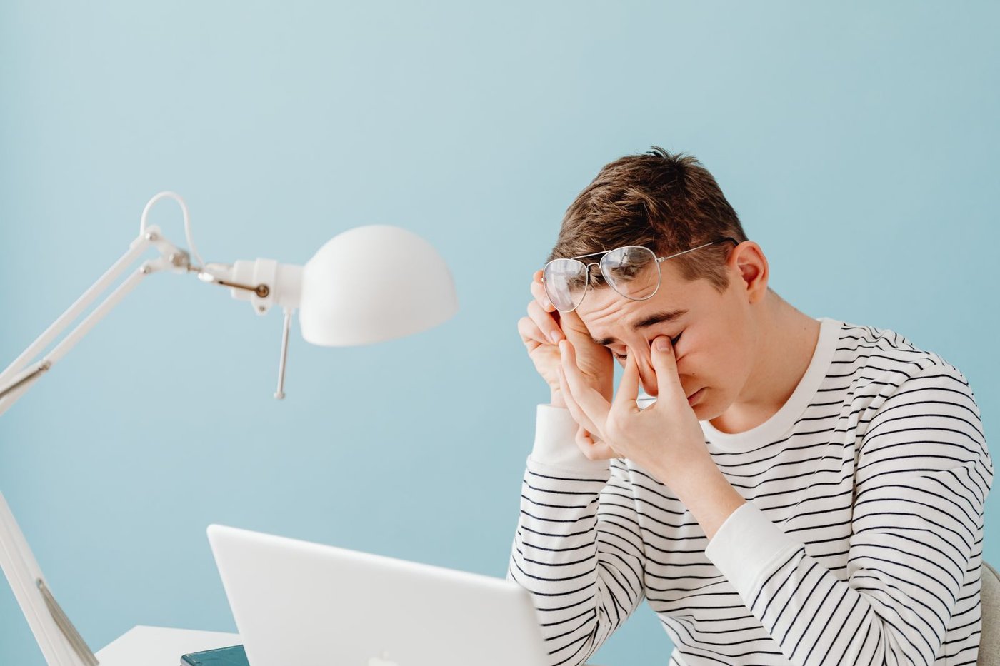 Young man rubbing his eyes at a desk after prolonged screen work, glasses pushed up on his forehead