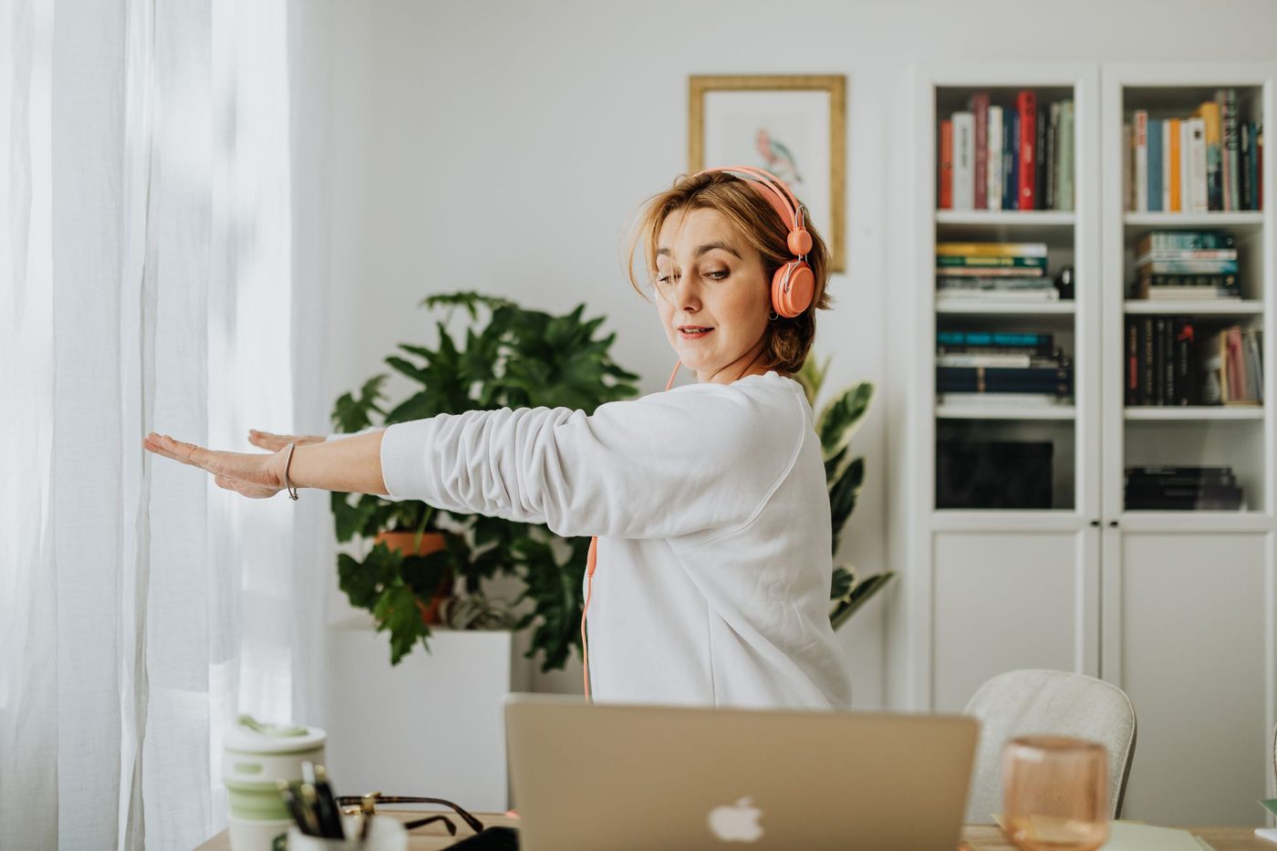 Woman in headphones standing at her home-office desk with arms stretched out to the side during a short stretch break