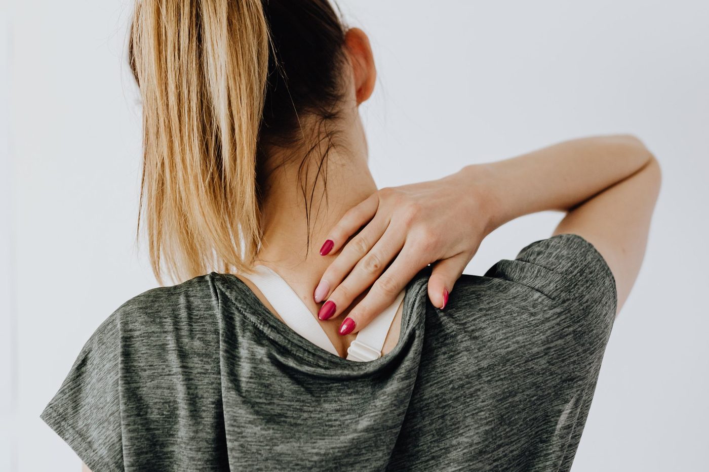 Back view of a woman in a gray T-shirt pressing a hand against the back of her neck and shoulder