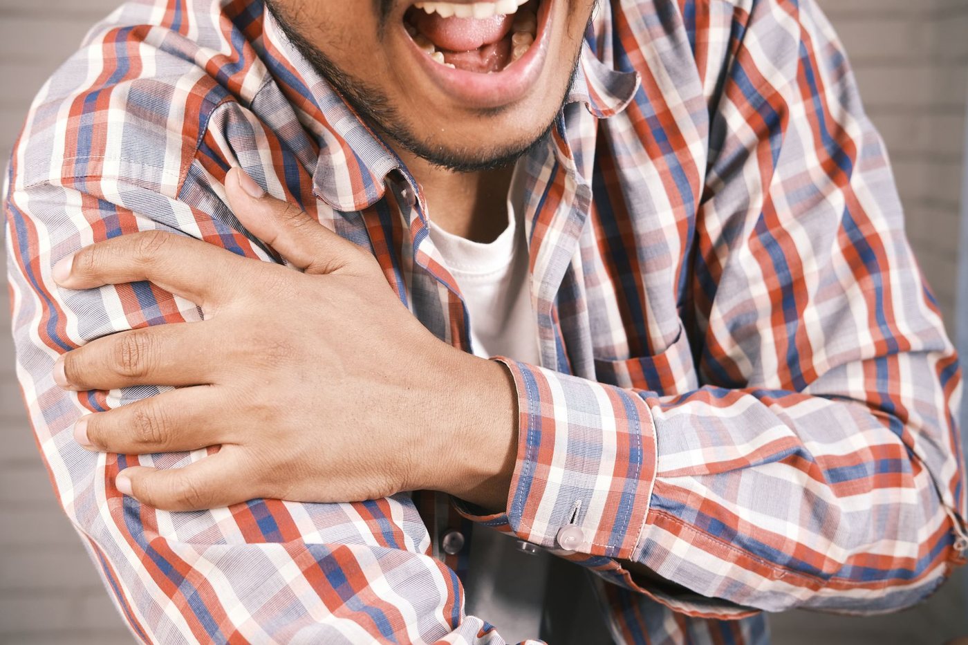 Close-up of a man in a plaid shirt gripping his right shoulder with a grimace of discomfort