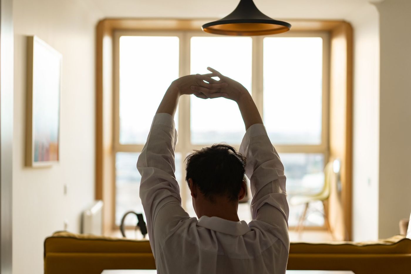 Man seen from behind stretching both arms overhead with hands clasped, in a sunny room