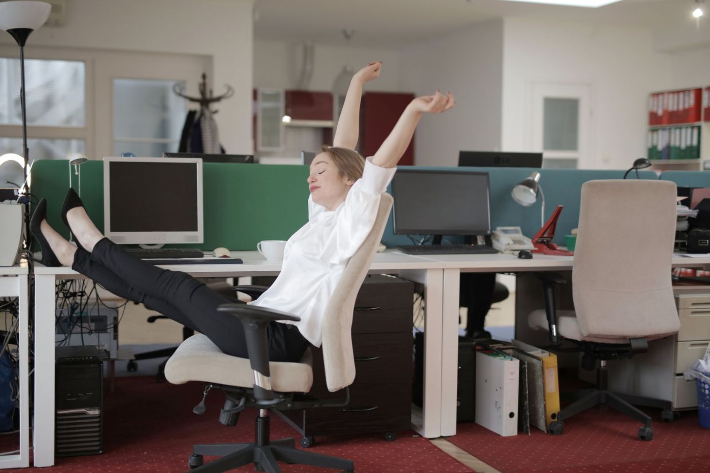 Office worker leaning back in a desk chair stretching both arms overhead in an open-plan office