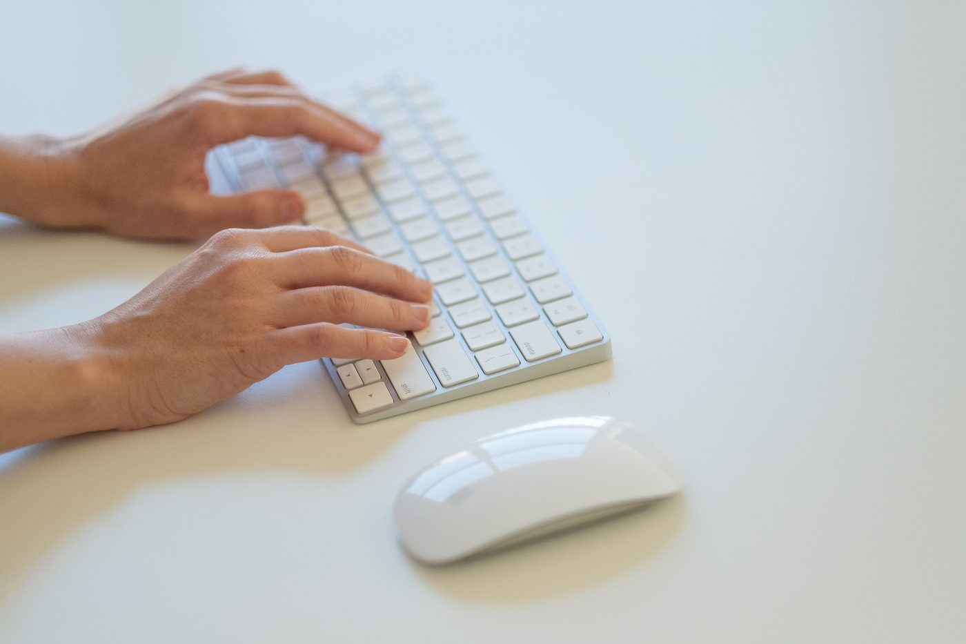 Hands typing on a white keyboard with a wireless mouse beside it on a light desk